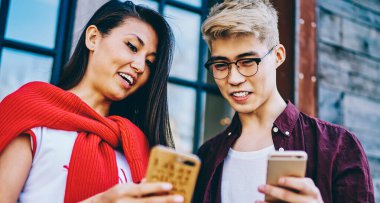 Neşeli hipster Asyalı öğrenciler ellerinde modern akıllı telefonlar ve çevrimiçi video izliyorlar şehir ortamında dikiliyorlar. Pozitif Japon arkadaşlar hücresel ağlarda tasarım profillerini tartışıyorlar.