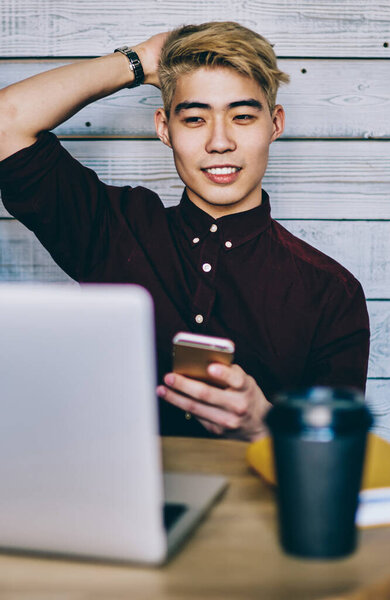 Asian hipster student holding smartphone in hand while resting and watching video on website on modern laptop connected to wireless internet.Chinese young man freelancer have break at netbook