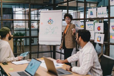 Genç, kendine güvenen bir kadın, iş arkadaşlarıyla, günlük kıyafetlerini giymiş, dizüstü bilgisayarla masaya oturmuş proje detaylarını tartışırken ve ofiste birlikte çalışırken çizelgeleri analiz ediyor.