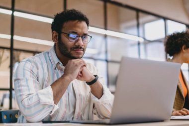 Ciddi İspanyol erkek kıvırcık saçlı, gündelik giysiler ve gözlüklerle masada dizüstü bilgisayar ve katlanmış ellerle modern çalışma alanında cam duvarla proje üzerinde çalışıyor.