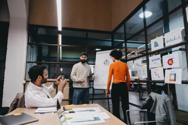 Bir grup çok ırklı iş arkadaşı gündelik giysiler içinde masanın etrafında kağıtlarla ve çizelgelerle dikilirken aynı zamanda cam duvarlı modern ofiste birlikte çalışıyorlar.