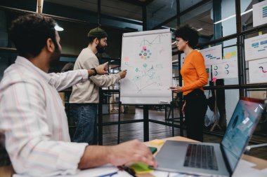 Gündelik giysiler içinde modern ofiste çizelgelerin yanında duran ve birlikte proje üzerinde çalışırken dizüstü bilgisayarla çizelgeleri analiz eden iş arkadaşlarının yan görüntüsü.