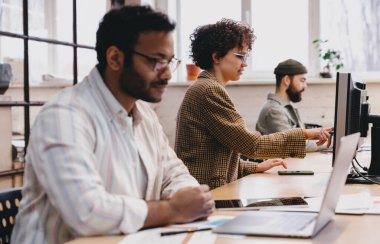 Yoğunlaştırılmış kadın işçi diğer iş arkadaşlarıyla birlikte gündelik giysiler içinde masamızda dizüstü bilgisayarla oturuyor ve modern çalışma alanı projesinde çalışırken bilgisayarın ekranına dokunuyor.