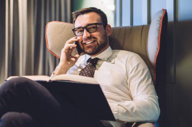 İş ortağıyla mobil sohbet sırasında koltukta oturan ve kameraya gülümseyen yakışıklı erkek tüccarın portresi, laptop cihazıyla kopyalama alanı ile eşzamanlı çalışma alanı