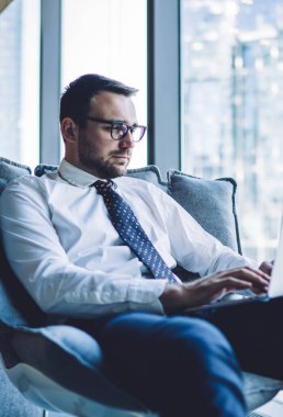 Gözlüklü yakışıklı adam dijital teknolojiyle online muhasebe yapıyor. Ofis kablosuz ağına bağlı. Modern dizüstü bilgisayarla belgelerle çalışan zeki bir erkek tüccar.