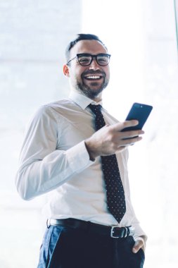 Ofis penceresinin yanında duran neşeli erkek muhasebecinin yarım boy portresi modern cep telefonu elinde ve iş molasında kameraya gülümsüyor, mutlu iş adamı içeride teknoloji kullanıyor.