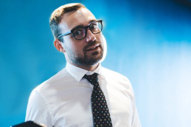 Görsel düzeltme için optik gözlüklü hayret içindeki işadamının yarım boy portresi akıllı telefon cihazını elinde tutuyor ve şirket ve ofis işlerinde çalışırken kameraya bakıyor.