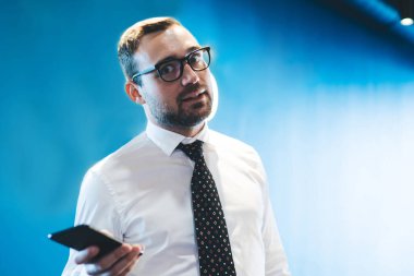 Görsel düzeltme için optik gözlüklü hayret içindeki işadamının yarım boy portresi akıllı telefon cihazını elinde tutuyor ve şirket ve ofis işlerinde çalışırken kameraya bakıyor.