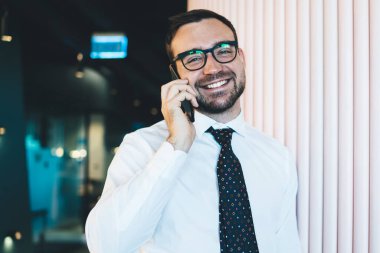 Gözlüklü, gülümseyen ve kameraya bakan neşeli bir adamın yarım boy portresi. Resmi beyaz gömlekli mutlu bir erkek muhasebeci. Cep telefonunda 4G kablosuz internet kullanıyor.