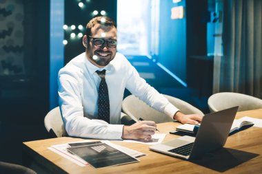 Optik gözlüklü mutlu işadamı, çalışma masasında oturup kağıt belgelerini analiz ediyor, bilgisayarında dijital iş yönetimi ve ortakların iletişimini inceliyor.