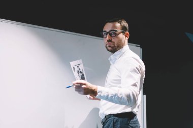 Klasik gözlüklü yakışıklı işadamının yarım boy portresi göz koruması sağlıyor evrak işlerini elinde tutuyor ve şirket tablosunun yanında poz veriyor.