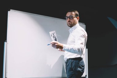 Klasik gözlüklü yakışıklı işadamının yarım boy portresi göz koruması sağlıyor evrak işlerini elinde tutuyor ve şirket tablosunun yanında poz veriyor.