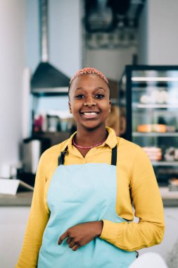 Modern, rahat bir kafede garsonluk yaparken neşeyle gülümseyen ve kameraya bakan önlüklü arkadaş canlısı Afrikalı Amerikalı kadın.