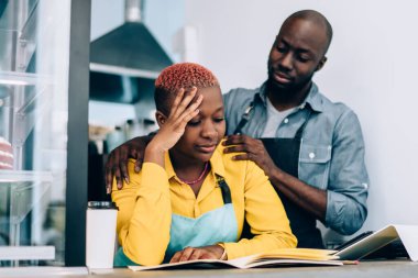 Önlüklü Afro-Amerikalı bir adam, küçük bir restoranda muhasebecilik yaparken yorgun bayan meslektaşını destekliyor ve rahatlatıyor.