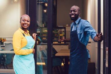 Kafe girişinin yanında durup müşterileri davet ederken önlüklü, gülümseyen ve kameraya bakan Afro-Amerikan erkek ve kadınların yan görüntüsü.