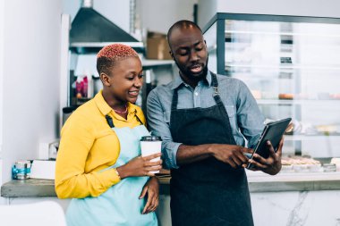 Önlüklü sakallı Afrikalı Amerikalı bir adam kafede çalışırken bir fincan sıcak içecekle modern tableti bayan meslektaşına gösteriyor.