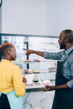 Önlüklü siyahi bir adam ve kadının birlikte kafede çalışırken pasta sergisinin yanında durup not defterine yazışlarının yan görüntüsü
