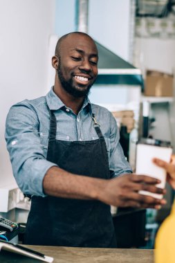 Önlüklü yakışıklı Afro-Amerikan adam gülümsüyor ve modern kafede çalışırken tanınmayan siyahi kadınlara bir fincan sıcak içecek veriyor.