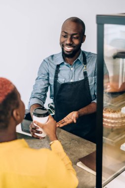 Yakışıklı, Afro-Amerikan erkek, gülümseyen ve bayan müşterilere sıcak bir şeyler ikram eden, rahat bir kafede çalışan.