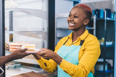 Dost canlısı Afrikalı Amerikalı bayan barmen neşeyle gülümsüyor ve küçük bir pastanede çalışırken siyah erkek müşteriye bir parça taze kek veriyor.