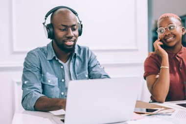 Neşeli Afrikalı Amerikalı kadın telefonda konuşuyor ve erkek meslektaşına bakıyor. Laptop kullanıyor ve kafede birlikte çalışırken müzik dinliyor.