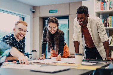 Çok ırklı genç erkek ve kadın, ofiste toplanırken proje ve kağıt üzerine yeni fikirler üzerinde çalışıyor.