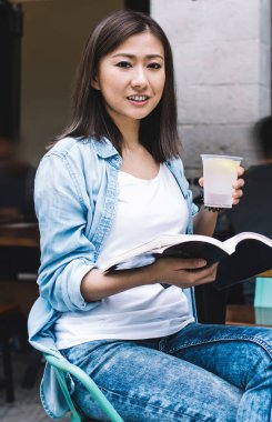 Esmer saçlı, edebiyat kitabı tutan çekici bir kadının yarım boy portresi. Boş zamanlarında dışarıda en çok satan kitapları okumak için kameraya bakan güzel bir kız öğrenci.