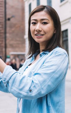 Portre pozitif Asyalı kadın kot gömlekli kameraya gülümseyen çevrimiçi video çekerken ve mobil uygulama ile sosyal ağlara paylaşırken, şehir tarihi merkezinde mutlu bir turist kaçamağı