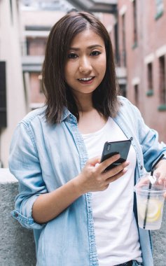 Uluslararası yolculuk sırasında şehir merkezinde soğuk limonlu içecek ile çekici Asyalı kadın turistin yarım boy portresi, internete bağlanan cep telefonlu çekici kadın.
