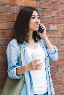 Kahveli çekici bir kız öğrenci cep telefonuyla cep telefonuyla sohbet etmeye gidiyor şehir merkezinde duruyor, cep telefonuyla konuşan genç bir kadın iyi gezen internete bağlanıyor.