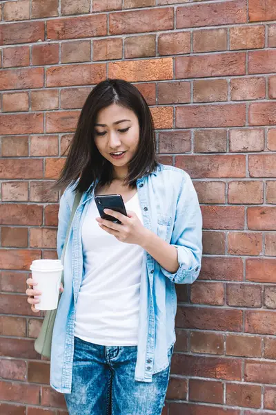 Modern cep telefonu cihazına sahip genç bir kadın şehir duvarının arka planında ve arama sitesinde dikilirken 4G internet bağlantısı kullanarak akıllı telefondan mesaj yazıyor.