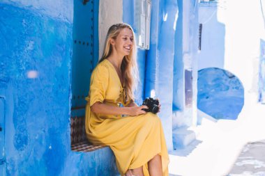 Eski kenti ziyaret etmek için Chaouen 'in mavi binasında dinlenirken geçmişe dönük ekipman kullanan pozitif kadın, açık havada çekim yapan neşeli Kafkasyalı turist