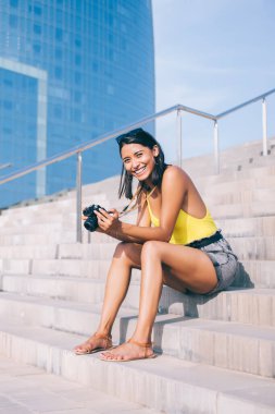 Sıradan giyinmiş heyecanlı hippi turist şehir setinde fotoğraf ekipmanlarıyla oturuyor ve boş zamanlarında kameraya gülümsüyor. Yüzünde güzel bir gülümseme olan neşeli İspanyol kadın.