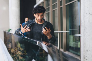 Modern binanın balkonunda duran yakışıklı, sıradan bir adam cep telefonunu şaşkınlıkla izliyor ve şok içinde el kol hareketi yapıyor.