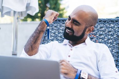 Modern dizüstü bilgisayarını kullanırken ve başarıyı el kaldırarak kutlarken, tatil köyünde sırtüstü uzanan sakallı mutlu etnik adam.