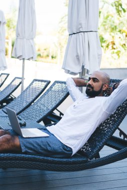 Güverte sandalyesinde dizlerinin üstüne çöküp el ele tutuşan ve terasta dinlenen mutlu etnik adamın yan görüntüsü.