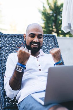 Neşeli etnik adam otelin terasında dizüstü bilgisayar izlerken ve başarıyı heyecanla kutlarken yumruklarını kaldırıyor.
