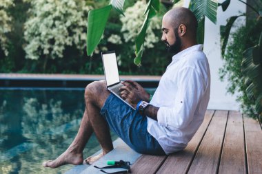 Gömlekli ve şortlu bir adamın bilardonun kenarında oturup uzaktan kumandayla tatil yapması.