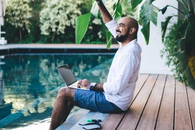 Havuzun kenarında oturmuş, iş gezisi yaparken el sallayan neşeli etnik adamın yan görüntüsü.