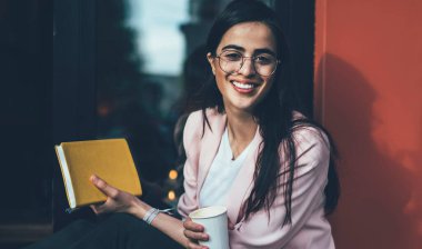 Kafenin yanında oturan, elinde kafein içeceği ve kameraya gülümseyen öğrenciler için web günlüğünün mutlu kadın yazarının portresi.