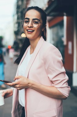 Gelişigüzel giyinen pozitif hippi kızın yarım boy portresi modern teknolojiyi telefon etmek ve kahve içmek için elinde tutarak şehir merkezini keşfederken kameraya gülümsüyor.