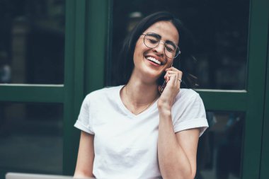 Görüş düzeltmesi için gözlüklü neşeli hippi kız şehir bölgesinde gezinirken eğlenceli telefon konuşmaları sırasında gülüyor, uygulamayla uluslararası telefon kavramı...