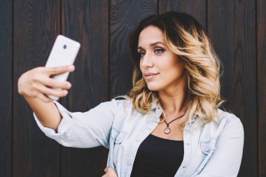 Çekici Milenyum hipster kız akıllı telefon web kamera üzerine selfie fotoğrafları tıklayarak ahşap arka plan yakın poz verirken, büyüleyici kafkas kadın sosyal ağda yayın için fotoğraf çekmek