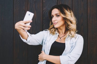 Çekici Milenyum hipster kız akıllı telefon web kamera üzerine selfie fotoğrafları tıklayarak ahşap arka plan yakın poz verirken, büyüleyici kafkas kadın sosyal ağda yayın için fotoğraf çekmek