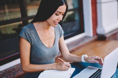 Modern bilgisayar ile kafeterya masasında otururken bilgi ders kitabında dergi için pozitif kadın gazeteci yazma makalesi, üniversite kursu çalışması için çekici kadın öğrenci notları yapma