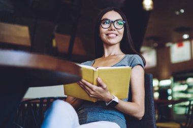 El kitabı ile neşeli hipster kız gülümseyen ve bir kenara bakarak kahve dükkanında edebiyat hobi için zaman harcama, vizyon düzeltme için gözlük mutlu kadın en çok satan tutan