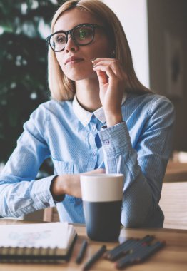 Not defteri ve kalemler ile ahşap masada otururken tasarım projesi için fikirler hakkında düşünme ciddi kadın öğrenci, kampüste gözler koruma öğrenme sağlamak için gözlükler içinde dalgın kadın