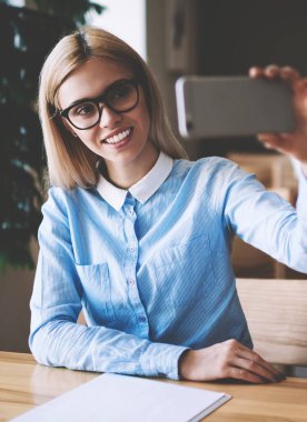 Üniversite sınıfı odasında öğrenme sırasında selfie fotoğraf tıklayarak için Web Kamera odaklanan gülümseyen kadın öğrenci, sosyal ağlarda paylaşmak için fotoğraf yapmak için akıllı telefon kullanarak pozitif kadın