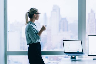 Gözlüklü, zarif, rahatlatıcı bir kadının bir fincan kahve içerken şehir manzarası olan büyük pencereye baktığı yan manzara.