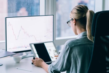 Pencerenin yanında tablet üzerinde çalışırken bilgisayar ekranında oturan gözlüklü modern iş kadınının arka planı